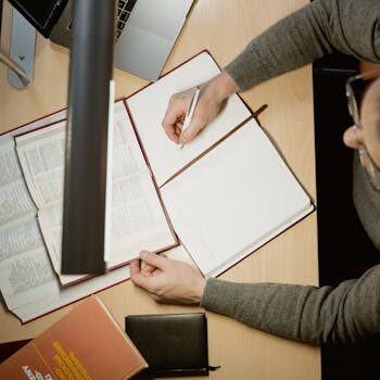 A person writing in a notebook while referencing books and a laptop, captured from above.
